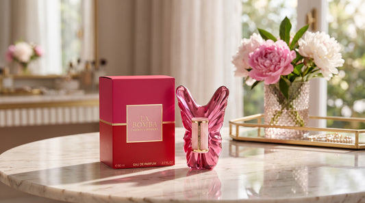 A pink butterfly-shaped perfume bottle stands next to its matching pink box on a marble table, with a blurred indoor setting and pink flowers in the background.