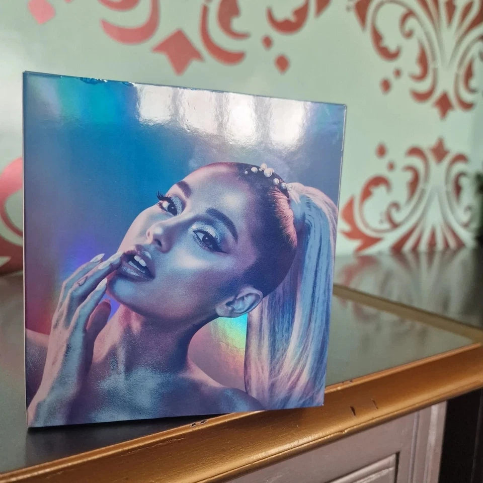 A square album or book-style cover featuring a stylized portrait of Ariana Grande with a holographic rainbow sheen, leaning on a wooden surface against a patterned wall.