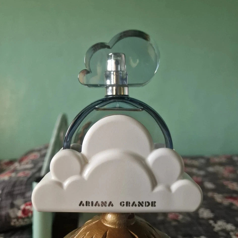 Ariana Grande perfume bottle with a white cloud-shaped base and a clear cloud-shaped cap, shown against a green wall and patterned bedspread.