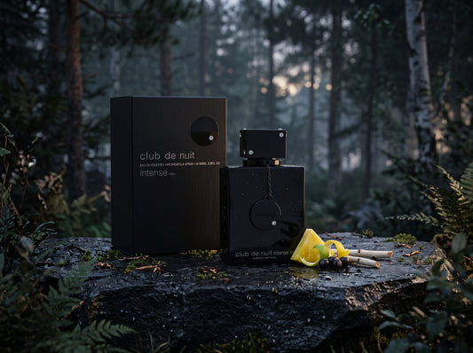 A black rectangular perfume box and matching bottle labeled 'club de nuit intense' sit on a wet log in a misty forest setting, with a yellow citrus slice and small props beside them.