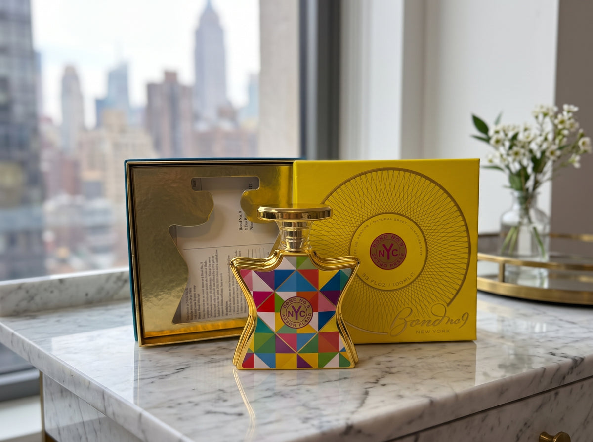A colorful geometric-patterned perfume bottle with gold detailing stands on a marble countertop in front of its yellow presentation box and inner packaging. A blurred city skyline is visible in the background near a window.