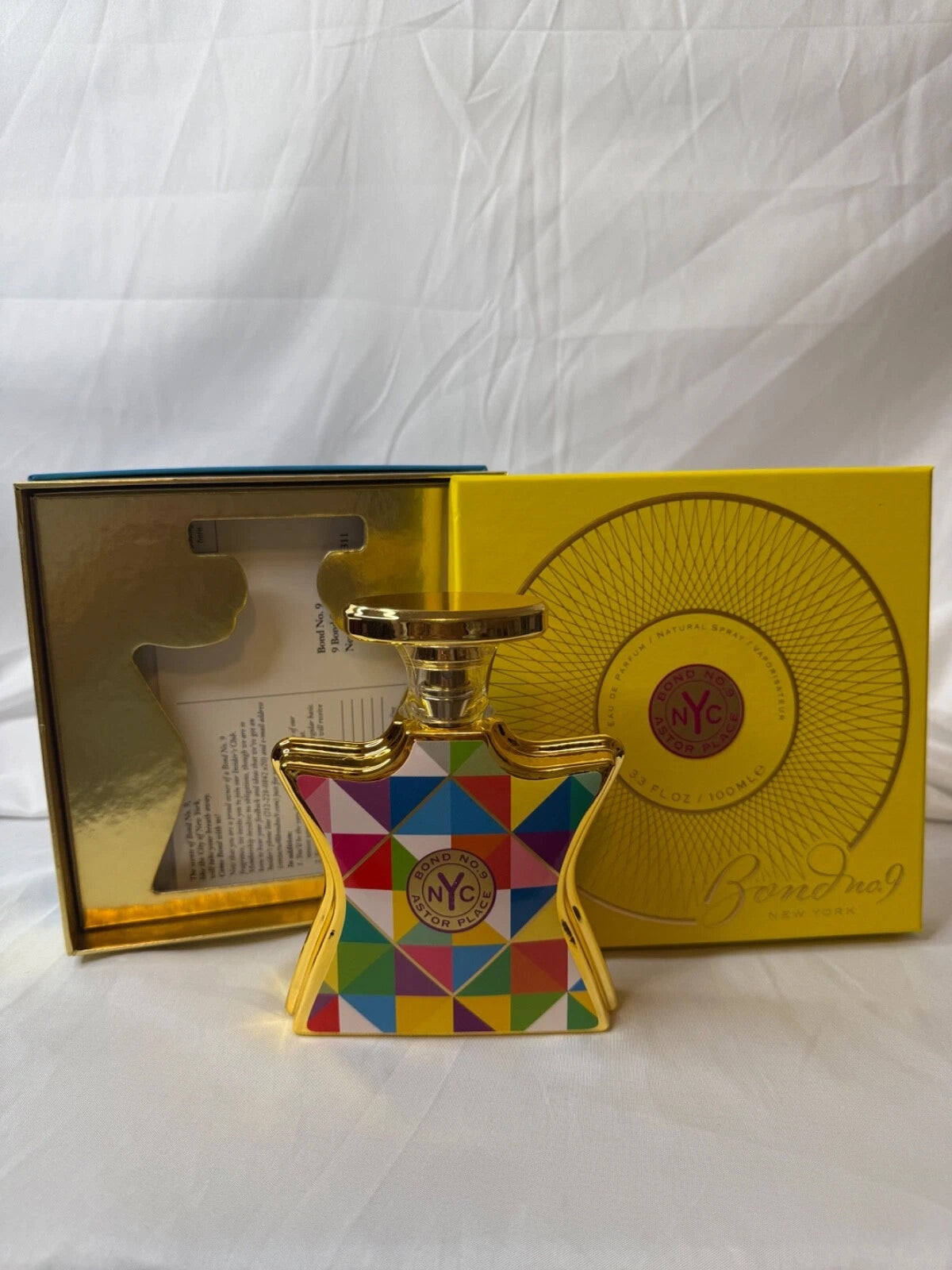Bond No. 9 perfume bottle with a colorful geometric pattern and gold cap, displayed in front of its yellow and gold presentation box on a white fabric background.