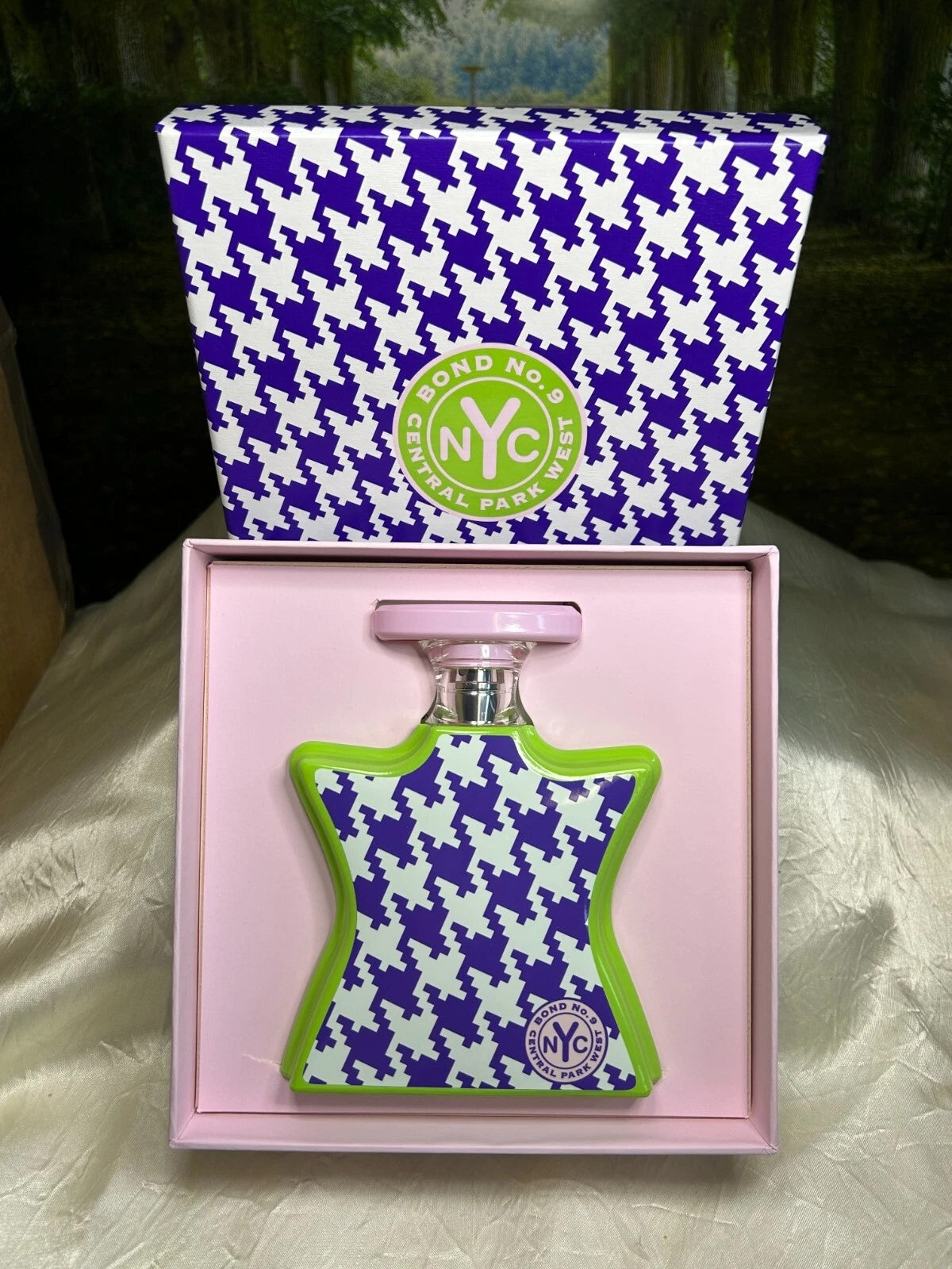 A perfume bottle with a purple and white houndstooth pattern and lime green trim, presented in a pink box with a matching branded box lid behind it.