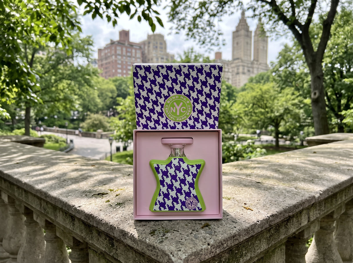 A perfume bottle in an open pink box with a matching patterned lid, placed on a stone railing outdoors with a blurred park and buildings background.