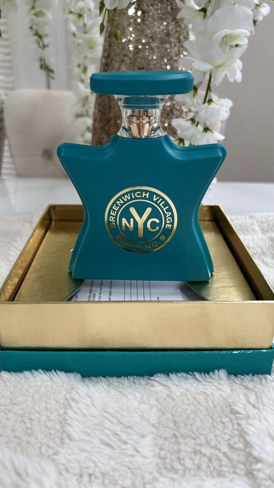 A teal green bottle with a gold circular 'Greenwich Village NYC' logo, displayed on a gold tray over a white fluffy surface with white flowers in the background.
