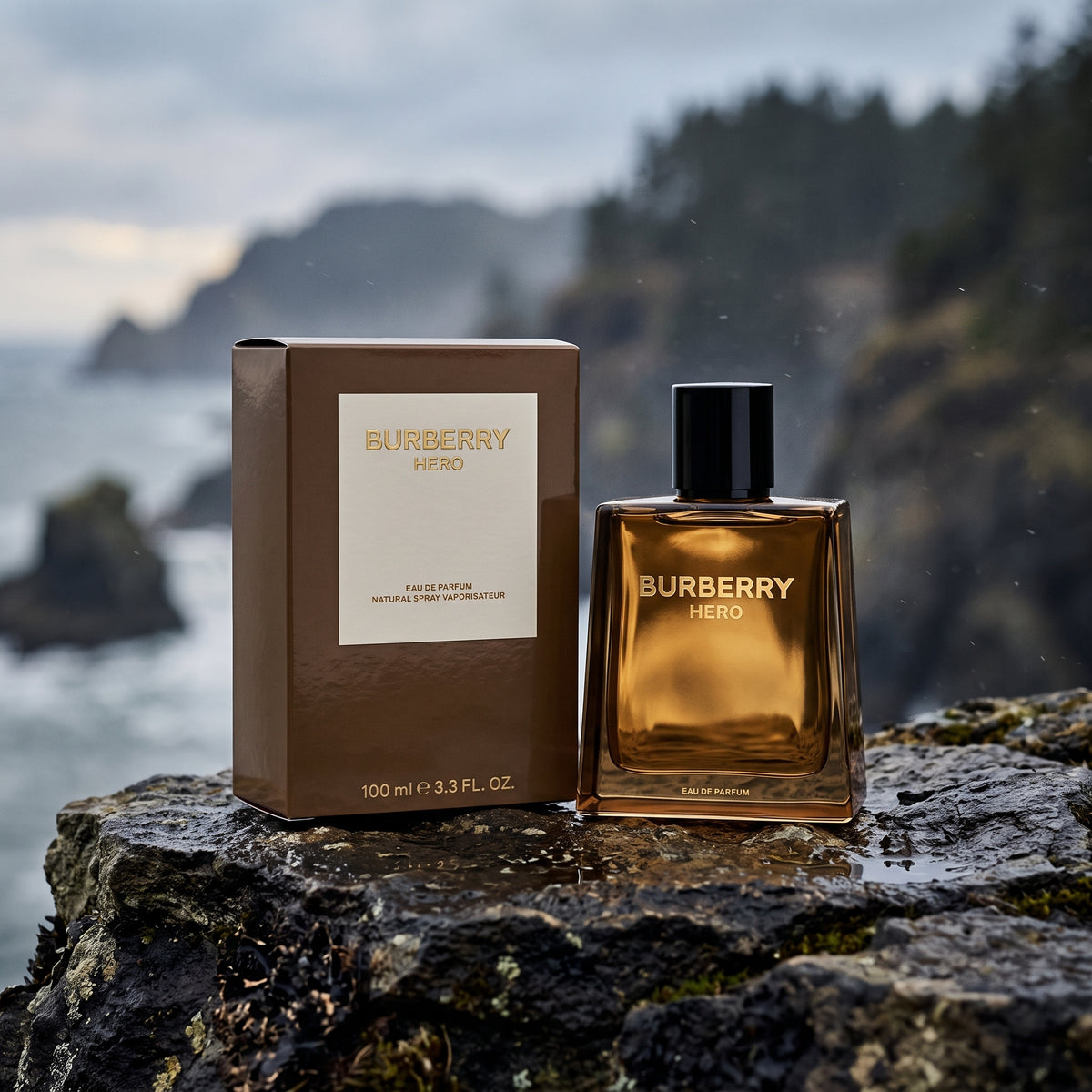 Burberry Hero Eau de Parfum bottle and its brown cardboard box placed on a wet dark rock with a blurred coastal cliff and ocean background.