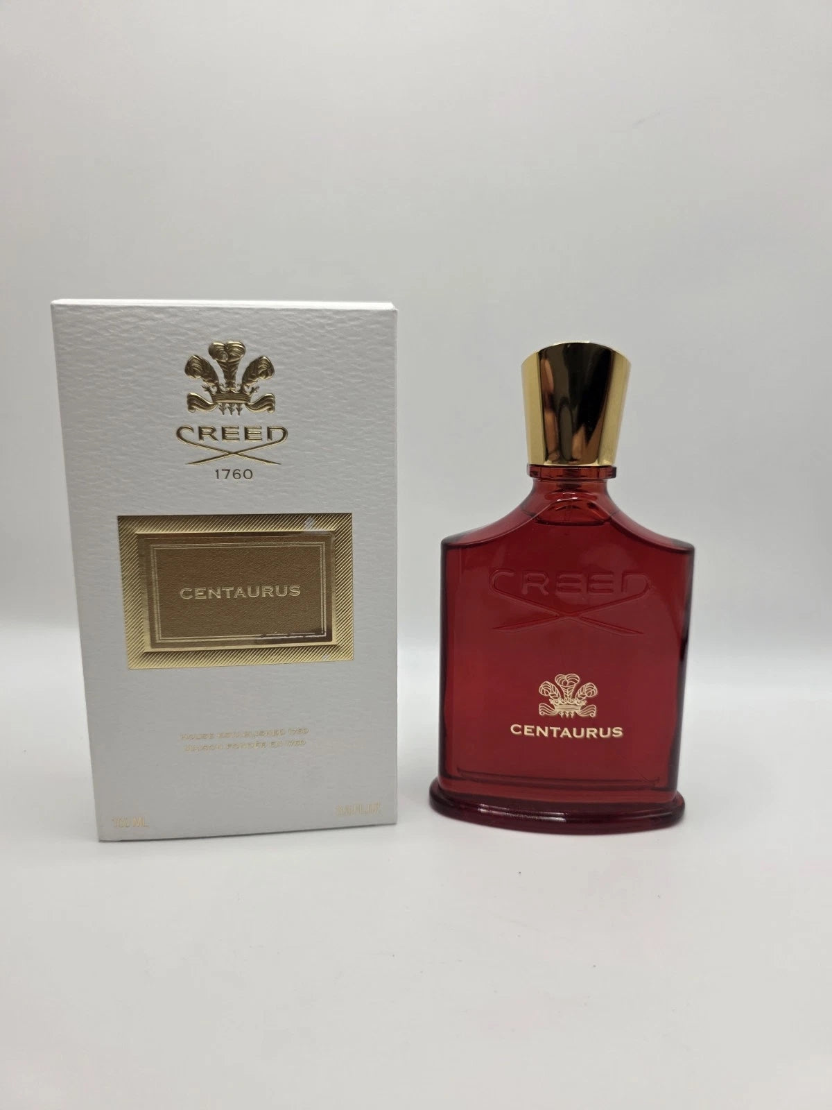 Creed Centaurus Eau de Toilette bottle in dark red glass with a gold cap, standing next to its white and gold retail box on a light gray background.