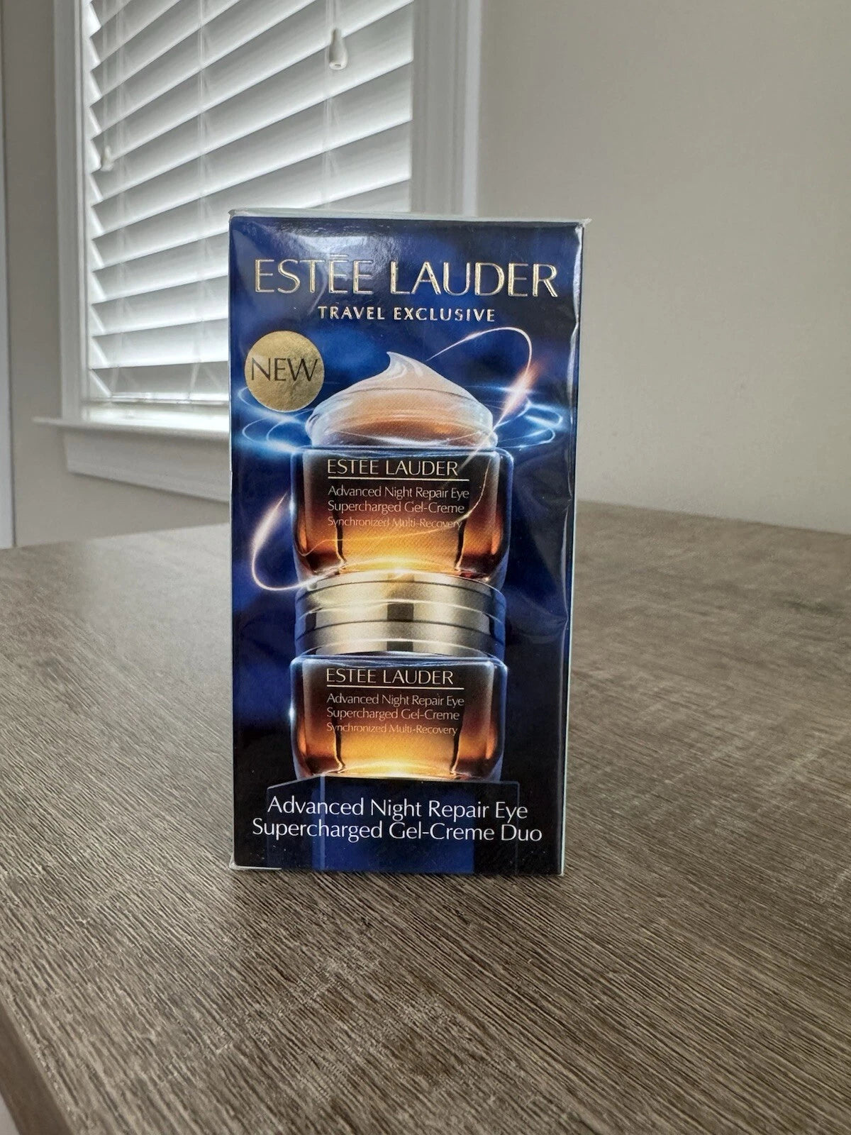 Estee Lauder Travel Exclusive Advanced Night Repair Eye Supercharged Gel-Creme Duo in its original blue and gold packaging, standing on a wooden table in front of a window with blinds.