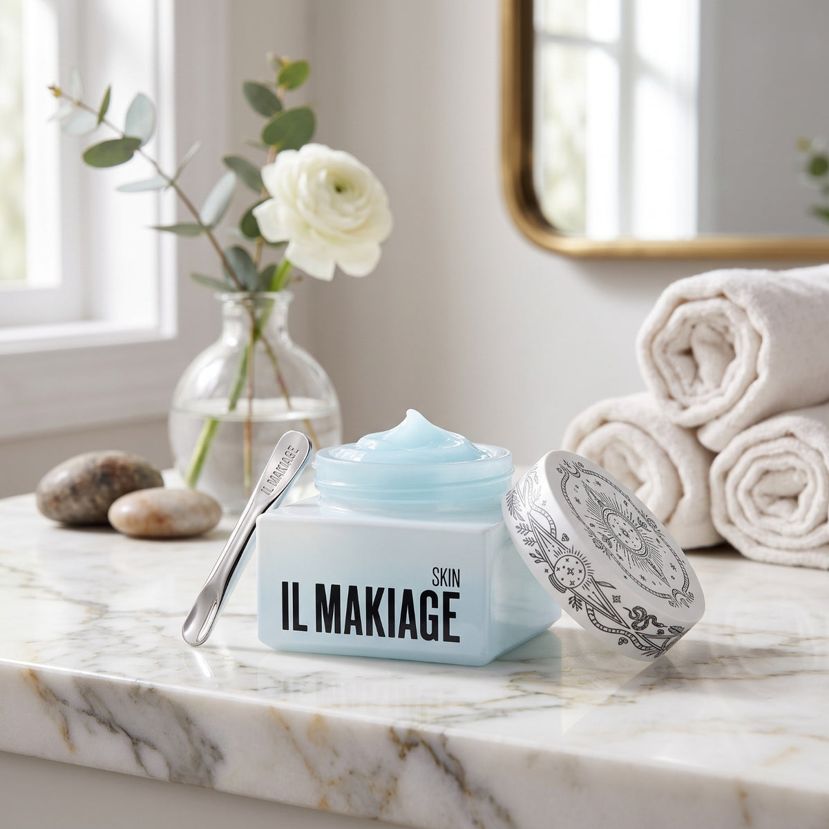 A cosmetic cream jar labeled 'IL MAKIAGE SKIN' sits open on a marble countertop with a spoon beside it. The scene includes rolled towels, a vase with a white rose, and a mirror in a bright bathroom setting.