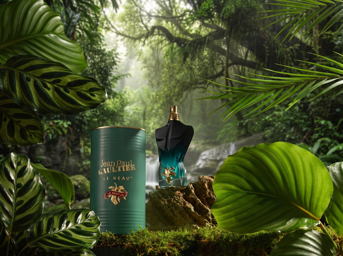Jean Paul Gaultier Le Beau perfume bottle and its signature teal cylindrical tin, styled in a lush, misty tropical jungle setting with green leaves framing the scene.
