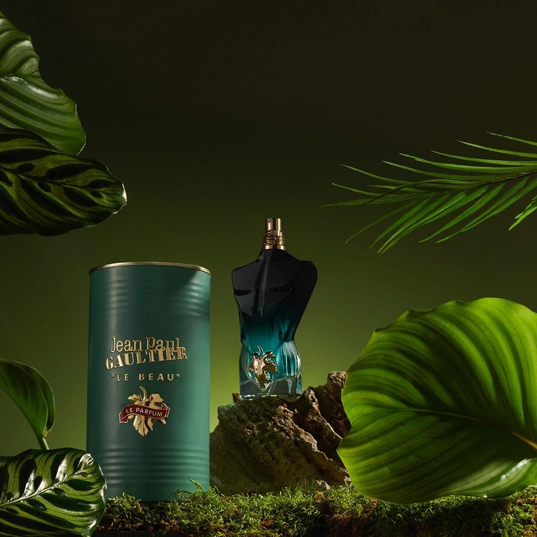 Jean Paul Gaultier Le Beau perfume bottle and its matching teal cylindrical packaging, styled with green tropical leaves on a mossy surface against a dark green background.