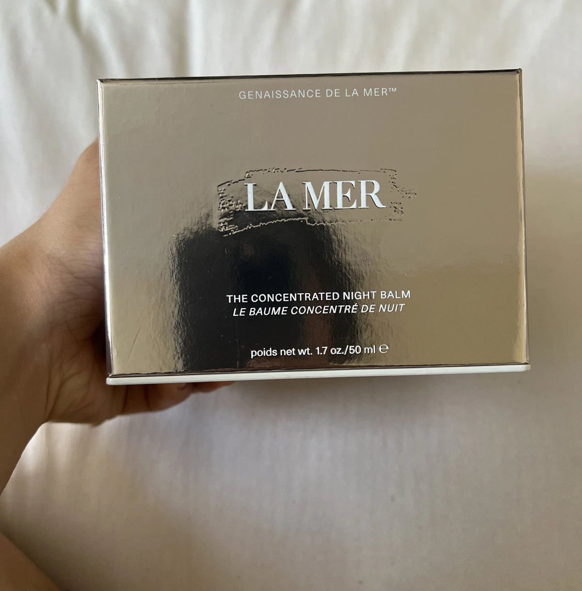 A hand holding a square metallic gold box of La Mer The Concentrated Night Balm against a soft beige fabric background.