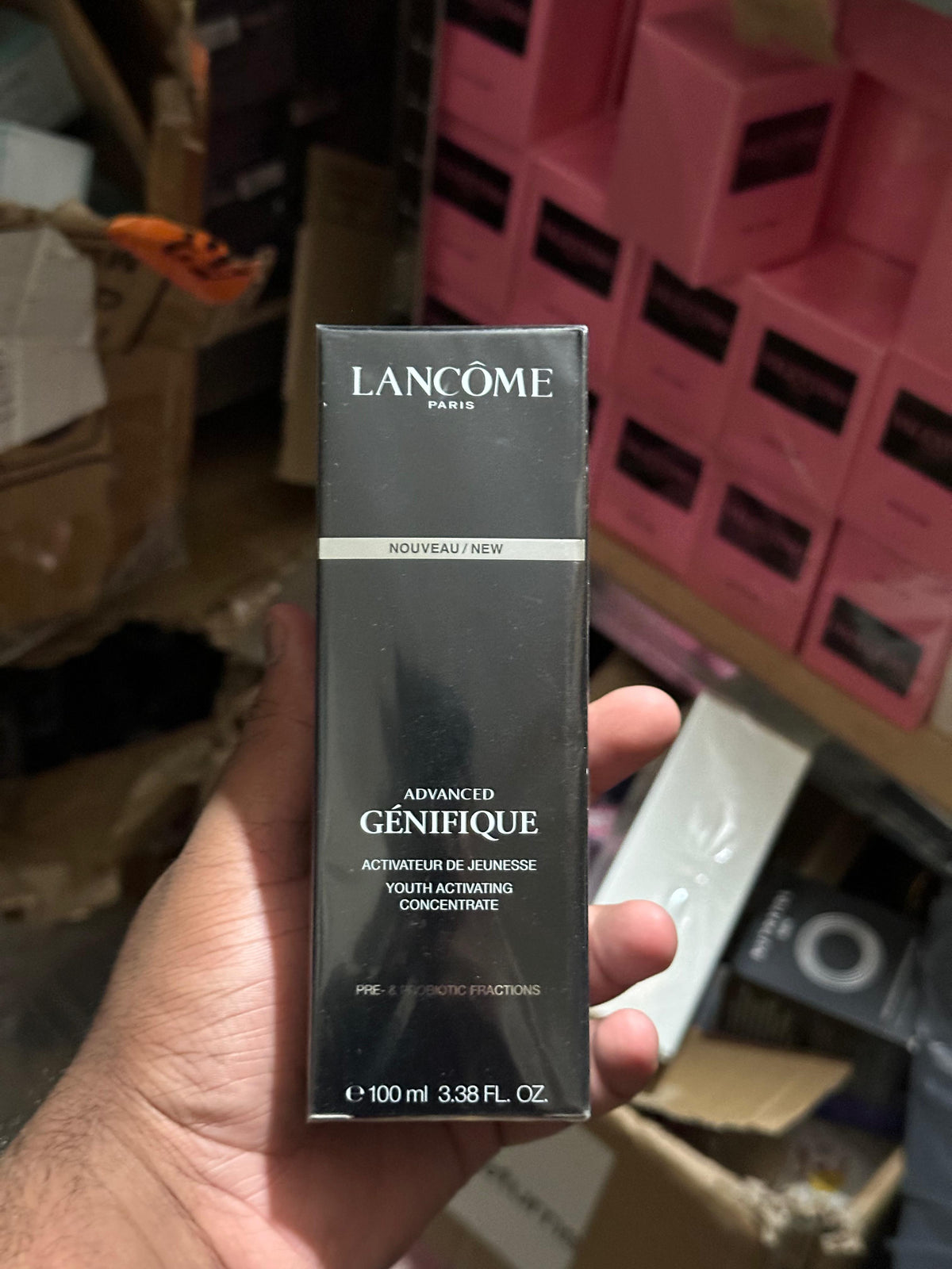 A hand holding a black Lancôme Paris Advanced Génifique Youth Activating Concentrate box, with a cluttered warehouse or store background filled with pink product boxes.