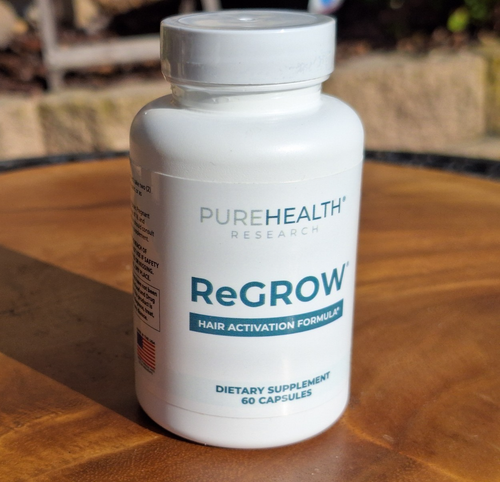 A white bottle of PureHealth Research ReGROW Hair Activation Formula dietary supplement, placed on a wooden surface with a blurred outdoor background.