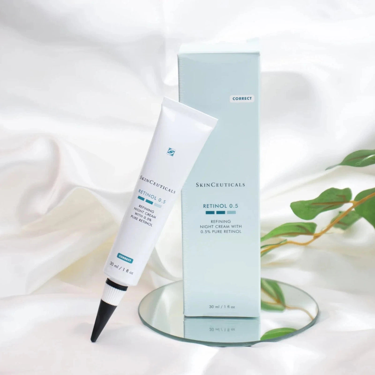 SkinCeuticals Retinol 0.5 retinoid cream shown with its light blue outer box and white squeeze tube, styled on a reflective glass plate with white draped fabric and green leaves in the background.