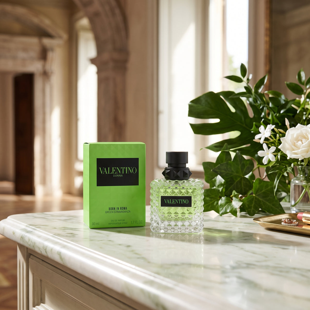 A bottle of Valentino 1991 & 2021 Eau de Parfum with a black cap sits on a white marble counter next to its signature lime green box, with a blurred background of an elegant interior and a floral arrangement.