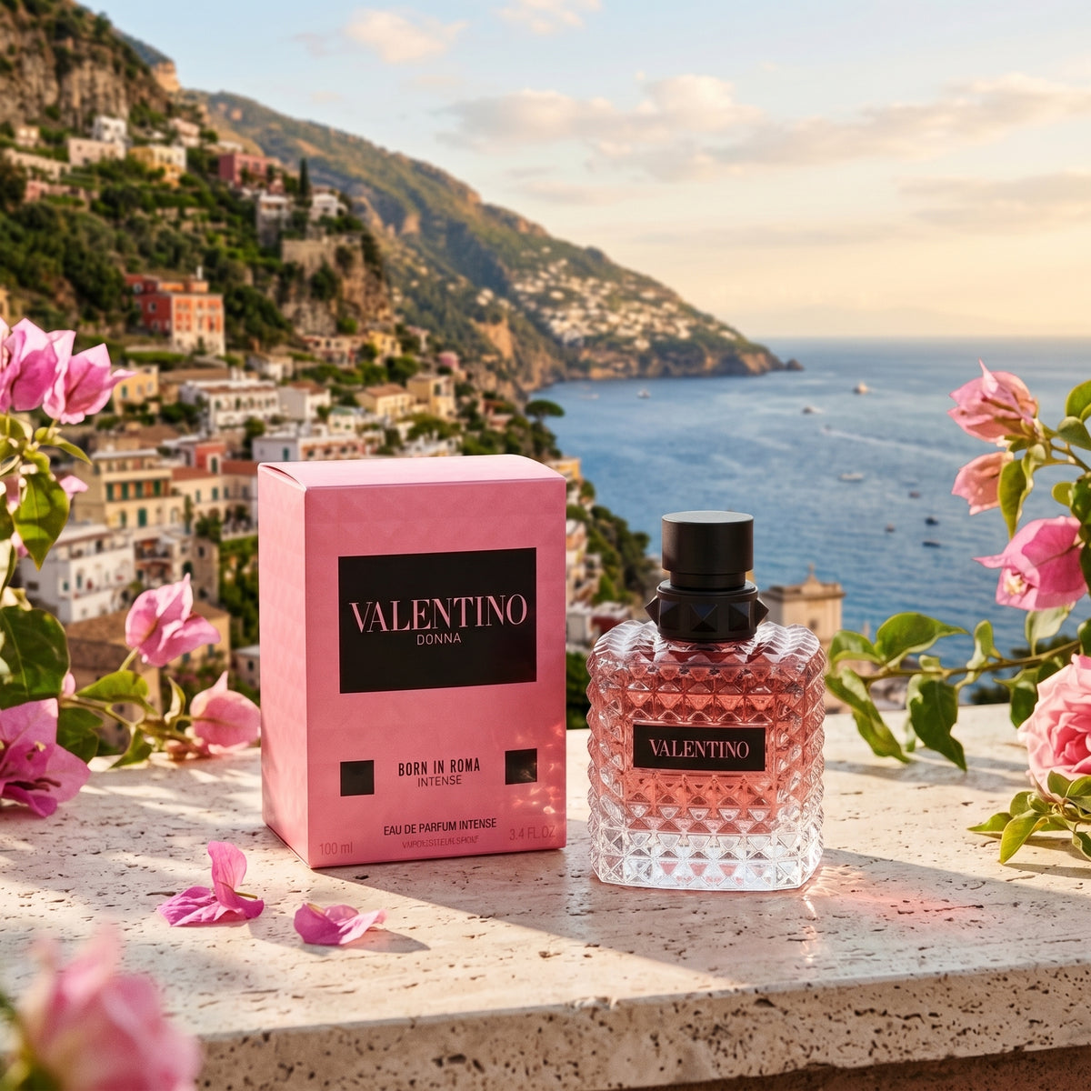 Valentino Born in Roma Eau de Parfum bottle and its pink box displayed on a stone ledge with pink flowers, set against a scenic background of the Amalfian coast and sea.