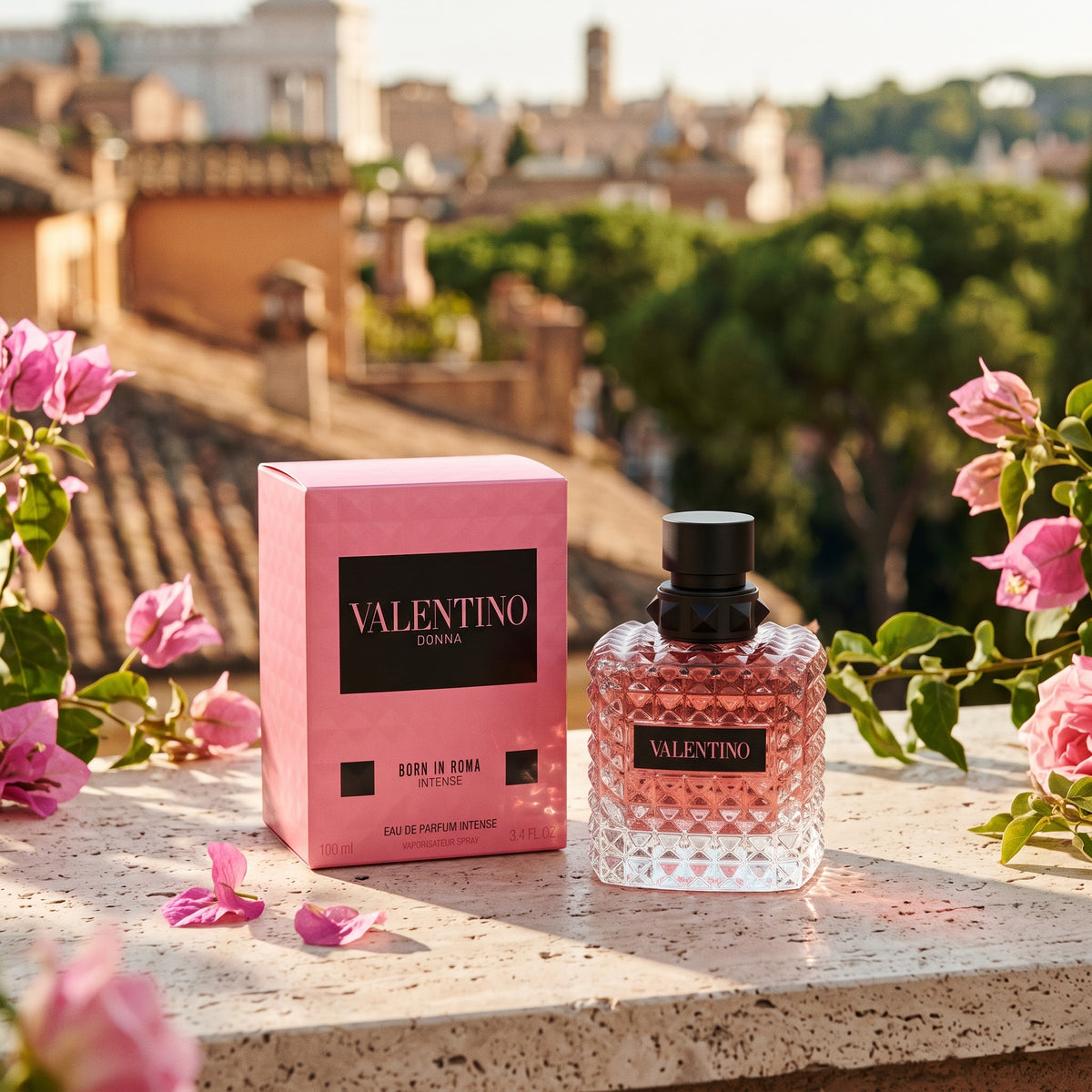 Valentino Donna Born in Roma Eau de Parfum bottle and its pink packaging box, styled on a stone ledge with pink flowers against a blurred background of Rome.