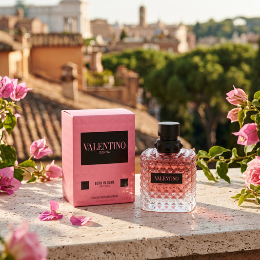 Valentino Donna Born in Roma Eau de Parfum bottle and its pink packaging box, styled on a stone ledge with pink flowers against a blurred background of Rome.