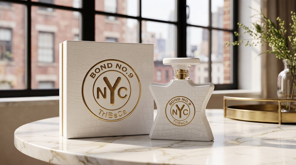 A star-shaped white perfume bottle and its matching square box with gold 'Bond No. 9 NYC TriBeCa' branding, displayed on a marble table in a bright room with a window and plants in the background.