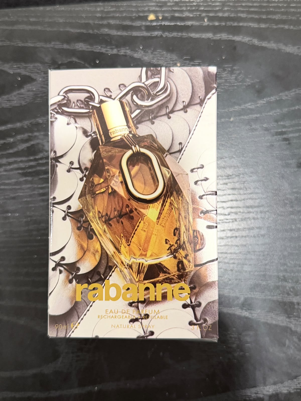 A boxed fragrance bottle labeled Rabanne Eau de Parfum placed on a dark wood surface. The box shows a faceted amber bottle with a gold cap and chain motif, and the brand name Rabanne in yellow text.