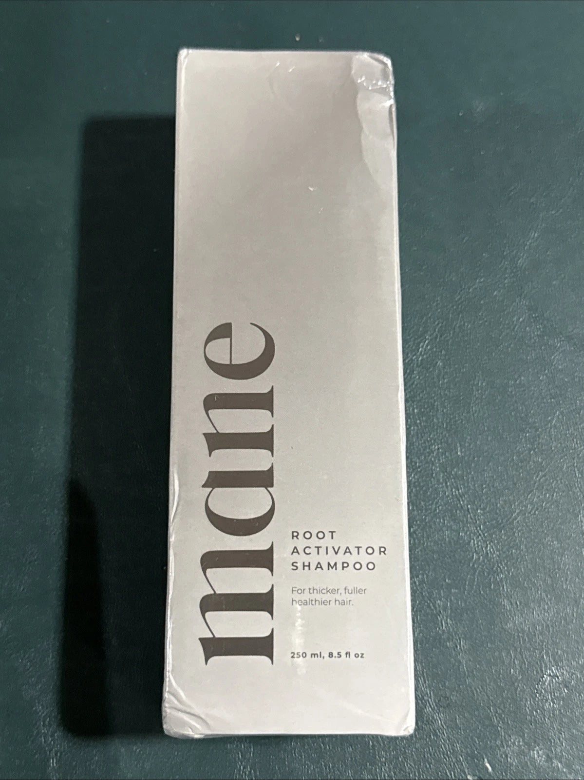A boxed bottle of mane Root Activator Shampoo, still sealed in its clear plastic wrap, standing upright on a dark teal surface.