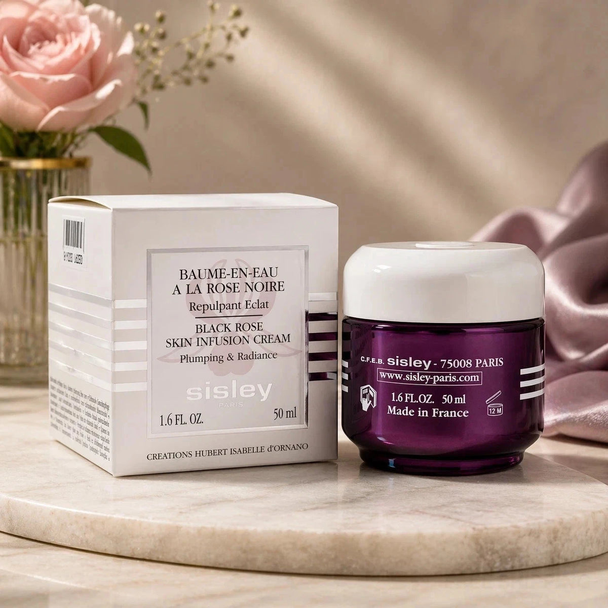 Sisley Black Rose Skin Infusion Cream 50ml jar next to its white packaging box, displayed on a marble surface with a pink rose and draped satin in the background.