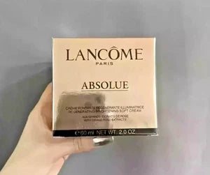 A hand holding a square box of Lancôme Paris Absolue Regenerating Brightening Soft Cream against a plain gray background.