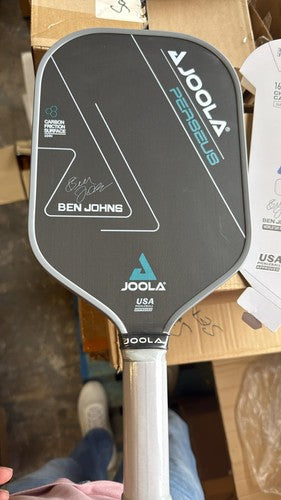 A signed Joola Pearls Ben Johng pickleball paddle, featuring a dark gray face with white and blue graphics, a silver handle, and an autograph, held in front of a cardboard box.